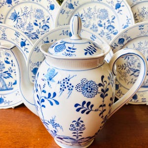 May include: White porcelain teapot and plates with blue floral patterns. The teapot has a lid and handle, and is surrounded by several matching plates. The design features detailed blue floral and leaf motifs, creating a classic, elegant aesthetic.