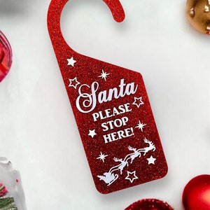 Op de afbeelding: Rode glitter deurhanger met een witte haak en de tekst "Santa Please Stop Here!" met een wit rendier en een slee.