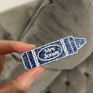 Pu&ograve; includere: Un'etichetta a forma di matita blu glitterata con contorno bianco e il testo "Mrs. Jones" in un ovale bianco.