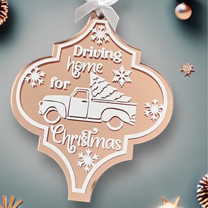 Puede incluir: Un adorno navideño de color oro rosa con forma de azulejo marroquí tradicional con un camión blanco que lleva un árbol de Navidad. El adorno dice "Driving home for Christmas".