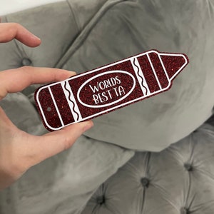 Pu&ograve; includere: Un cartello rosso a forma di matita glitterata con contorno bianco e il testo "Worlds Best Ta" in un cerchio bianco.