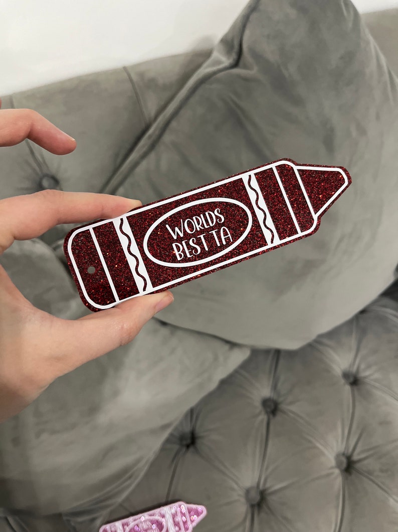 Pu&ograve; includere: Un cartello rosso a forma di matita glitterata con contorno bianco e il testo "Worlds Best Ta" in un cerchio bianco.
