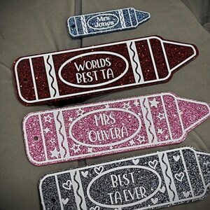 Pu&ograve; includere: Quattro cartelli a forma di matita ricoperti di glitter con testo bianco. I cartelli recitano "Mrs. Jones", "Worlds Best TA", "Mrs. Olivera" e "Best TA Ever".
