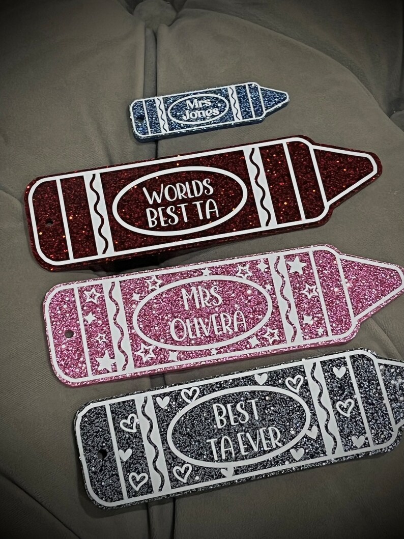 Pu&ograve; includere: Quattro cartelli a forma di matita ricoperti di glitter con testo bianco. I cartelli recitano "Mrs. Jones", "Worlds Best TA", "Mrs. Olivera" e "Best TA Ever".
