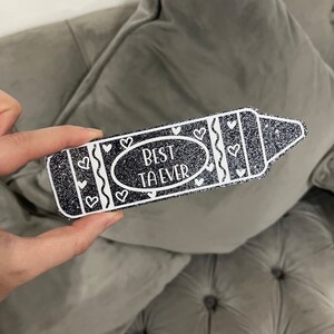 Pu&ograve; includere: Un pastello nero glitterato con contorno bianco e cuori. Il pastello dice "Miglior Insegnante di Sempre" in testo bianco.