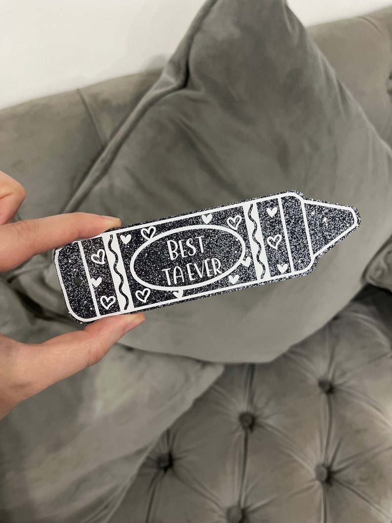 Pu&ograve; includere: Un pastello nero glitterato con contorno bianco e cuori. Il pastello dice "Miglior Insegnante di Sempre" in testo bianco.
