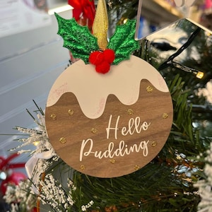 Puede incluir: Un adorno navideño de madera con forma de pudín navideño con un glaseado blanco en la parte superior y un fondo marrón. Tiene detalles de purpurina dorada y el texto "Hello Pudding" escrito en blanco. El adorno está decorado con una rama de acebo verde y bayas rojas.