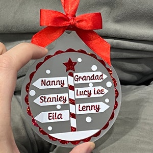 May include: A gray and white Christmas ornament with a red glitter finish. The ornament features a red and white candy cane pole with signs that read "Nanny", "Grandad", "Stanley", "Eila", "Lucy Lee", and "Lenny".