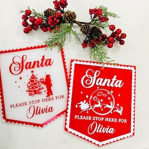 Könnte beinhalten: Zwei rote und weiße Schilder mit gewellten Rändern. Die Schilder sagen "Santa Please Stop Here For Olivia" mit einem Weihnachtsbaum und dem Weihnachtsmann auf dem einen Schild und einem Mond und Rentieren auf dem anderen.