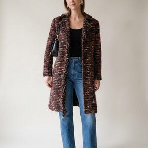 May include: A long-sleeved, knee-length tweed coat featuring a multi-colored textured weave in shades of black, brown, and rust with frayed edges. The coat is worn over a black tank top, blue denim jeans with raw hems, and dark red ballet flats.
