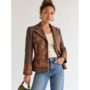 May include: A brown blazer featuring intricate orange, red, and gold floral and paisley embroidery along the front panels and sleeve cuffs. The blazer has a notched lapel, long sleeves, and is worn over a cream-colored top and blue denim jeans.