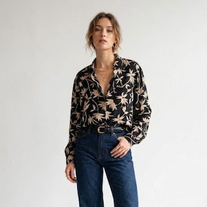 May include: A long-sleeved button-down shirt featuring a black base with a repeating beige botanical leaf pattern. The shirt is paired with dark blue denim jeans that have a black leather belt with a silver buckle at the waistline.