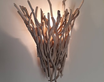 Puede incluir: Una lámpara de pared hecha de madera flotante natural. La lámpara está compuesta por numerosas ramas de madera clara y desgastada dispuestas en forma cónica, con una luz cálida que emana del interior. La lámpara está montada en una pared blanca.