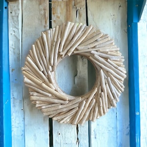 May include: A wreath made of driftwood branches, arranged in a circular shape. The wreath is a light brown color and has a natural, rustic look. It is hanging on a white wooden wall.