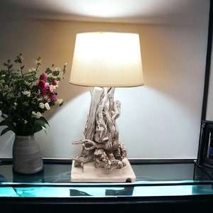 Op de afbeelding: Een witte drijfhouten tafellamp met een beige lampenkap. De lamp staat op een zwart oppervlak met een vaas met roze bloemen op de achtergrond.