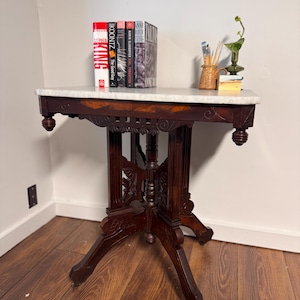 May include: An antique, dark wood side table with a white marble top. The table features ornate carvings and a decorative apron. Books, a small vase, and art supplies are displayed on the table. The table is approximately 30 inches tall.