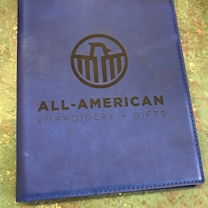 以下が含まれることがあります： 「ALL-AMERICAN EMBROIDERY + GIFTS」の文字と、鷲をあしらった円形ロゴが入った青いノート。ステッチの縁取りと、革のような質感の表面が特徴です。
