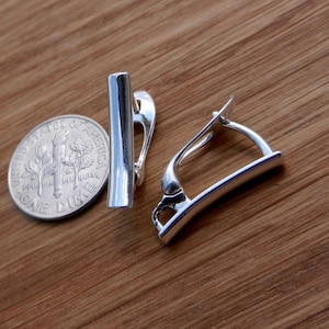 Puede incluir: Un par de pendientes de clip plateados. Los pendientes están hechos de un metal liso y curvado y tienen un diseño simple y moderno.