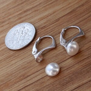May include: A pair of silver hoop earrings with a white pearl dangling from each hoop. The earrings are shown next to a US dime for size comparison.