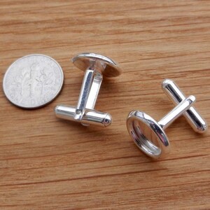 May include: Two silver cufflinks with round, empty, silver bezels. The cufflinks are on a wooden surface.