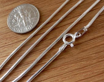 Puede incluir: Primer plano de un collar de cadena de serpiente plateada con cierre de mosquetón. Una moneda de diez centavos estadounidense está junto a la cadena para comparar el tamaño. La cadena tiene una superficie reflectante y texturizada.