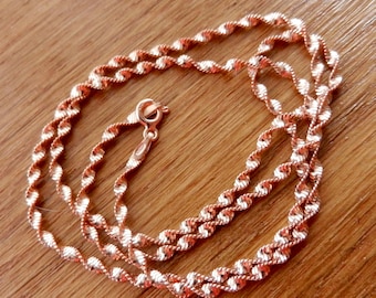 May include: A rose gold necklace with a twisted rope design. The chain is displayed on a wooden surface. The necklace has a delicate, textured appearance and a lobster clasp.