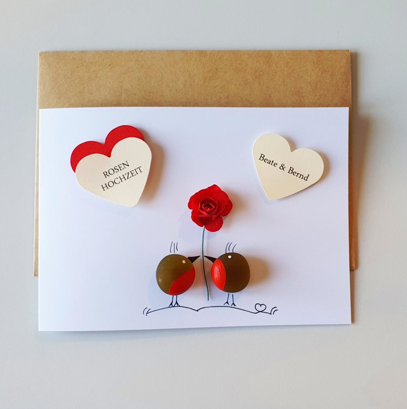 Op de afbeelding: Een witte kaart met een rode roos, twee rode vogels en twee witte harten. De harten hebben de tekst "Rosen Hochzeit" en "Beate & Bernd".