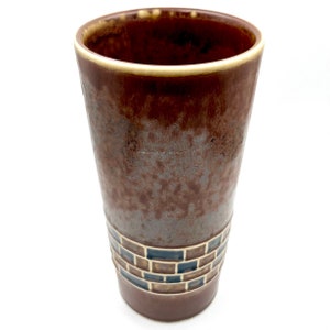 Puede incluir: Un jarrón de cerámica marrón con un patrón geométrico alrededor del centro. El jarrón tiene un esmalte marrón moteado con un borde marrón más claro.