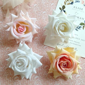 May include: Four artificial roses in different shades of pink, white, and peach. The roses are arranged on a pink background with a white pattern.