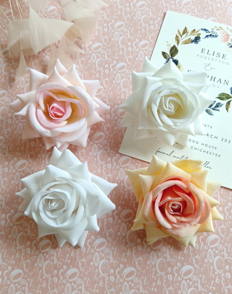 May include: Four artificial roses in different shades of pink, white, and peach. The roses are arranged on a pink background with a white pattern.
