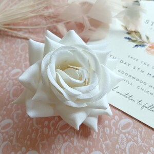 May include: A single white rose with a delicate, layered texture. The rose is on a pink background with a white pattern.