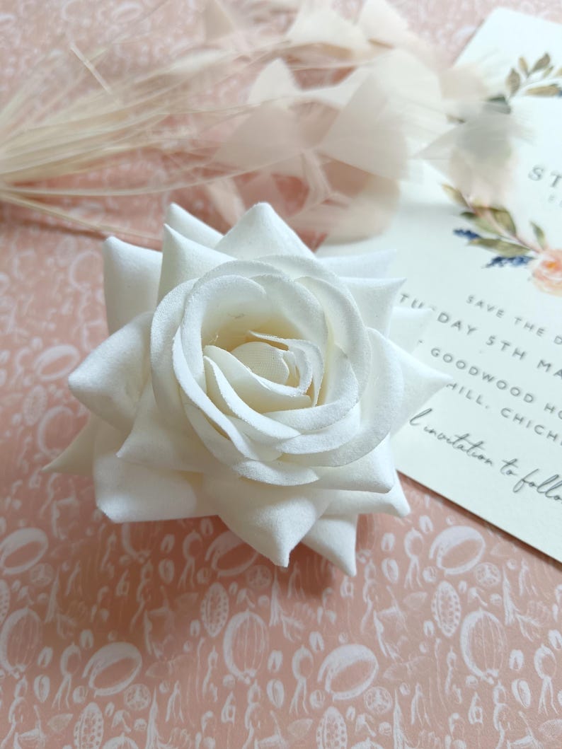 May include: A single white rose with a delicate, layered texture. The rose is on a pink background with a white pattern.