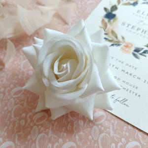 May include: A single white rose with a light pink background. The rose is a silk flower and has a delicate, realistic appearance.