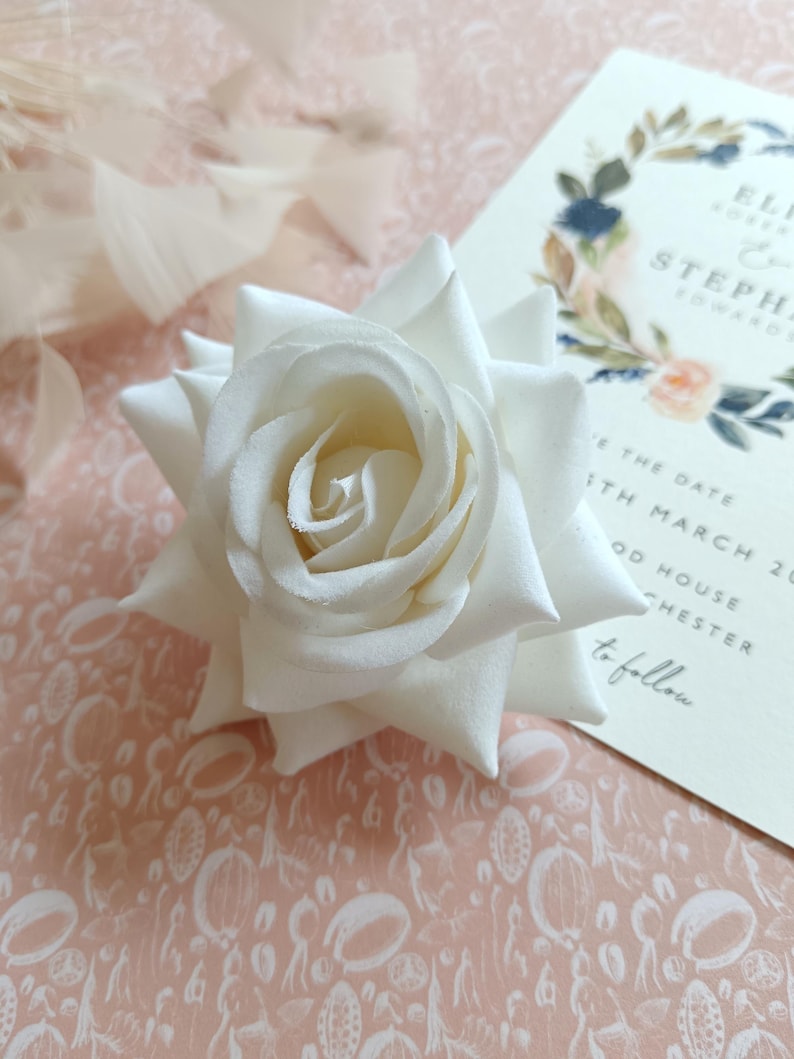 May include: A single white rose with a light pink background. The rose is a silk flower and has a delicate, realistic appearance.