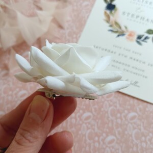 May include: A white fabric rose flower with a silver pin on the back. The rose is held in a hand.
