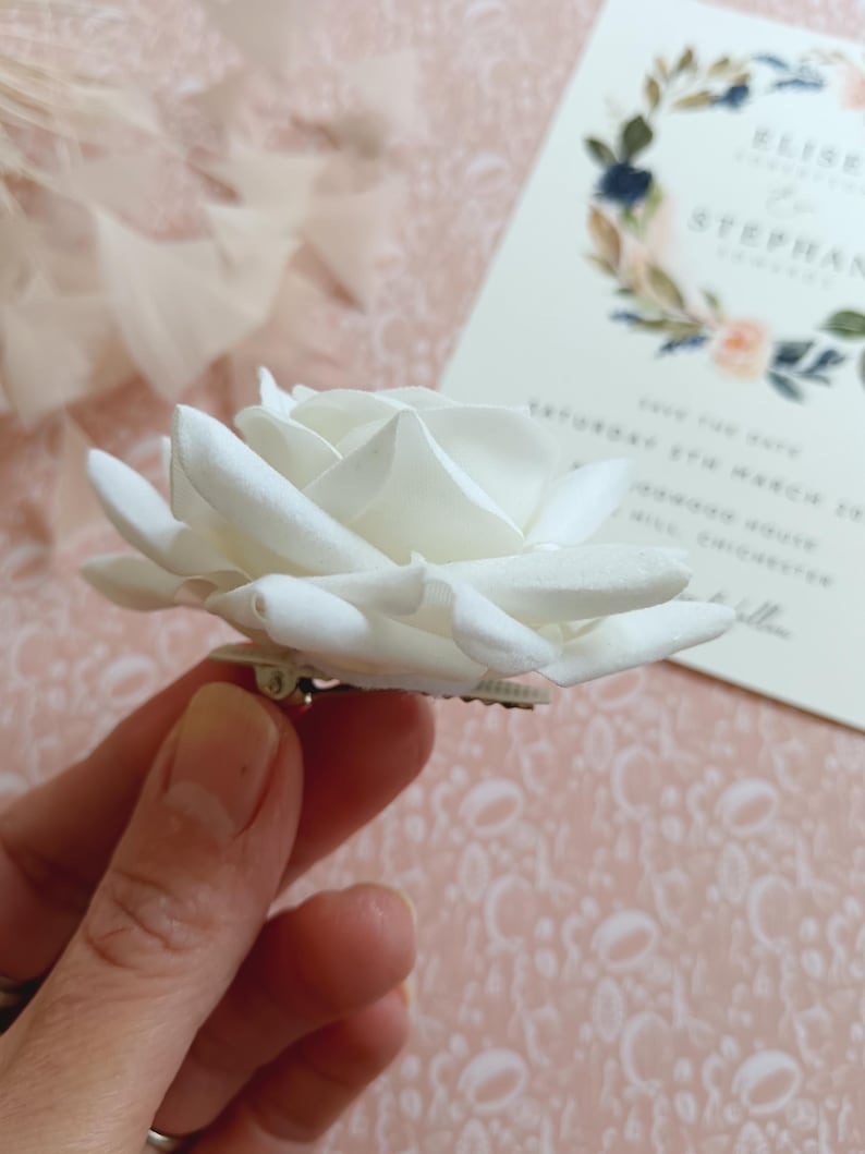 May include: A white fabric rose flower with a silver pin on the back. The rose is held in a hand.