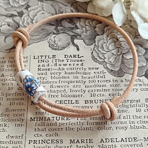 May include: A cream leather cord bracelet with a white ceramic bead decorated with a blue floral pattern. The bracelet is displayed on a vintage paper background with floral illustrations. The cord is knotted.