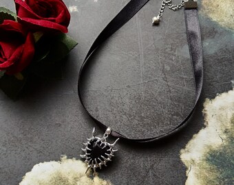 May include: A black choker necklace with a silver pendant. The pendant has a black center stone and a spiky silver frame. The necklace is made of black satin ribbon and has a silver chain clasp. Two red roses are in the background.