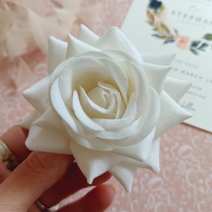 May include: A single white artificial rose with a layered, ruffled design. The rose is held in a hand with a ring on the finger.