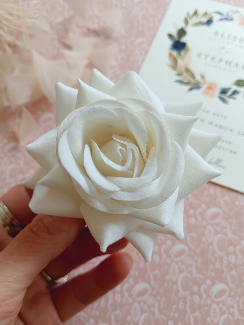 May include: A single white artificial rose with a layered, ruffled design. The rose is held in a hand with a ring on the finger.