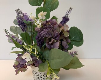 May include: A small potted floral arrangement with purple and white flowers and green leaves. The pot is white with black polka dots.