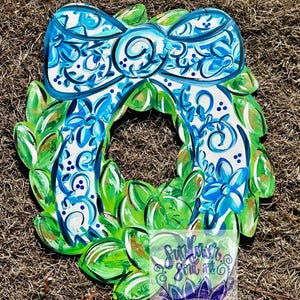 May include: A blue and white patterned bow with green leaves painted on a wooden wreath. The bow has a white background with blue swirls and flowers. The leaves are a bright green with gold accents.