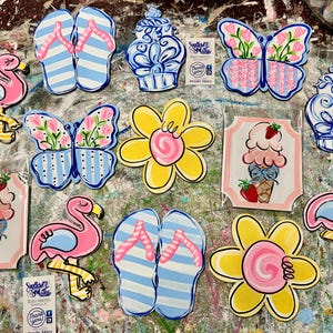 May include: A collection of hand-painted wooden cutouts featuring various designs including pink flamingos, blue and white striped flip-flops, blue and white ginger jars, colorful butterflies, yellow flowers, and ice cream cones with strawberries.