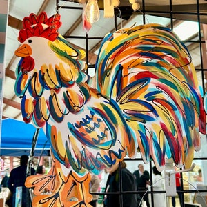 May include: A colorful rooster cutout with a white background and painted in shades of red, orange, yellow, blue, and green. The rooster is hanging on a black wire grid.