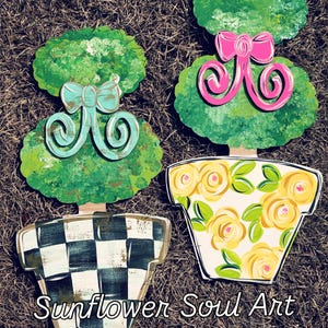 May include: Two painted wooden topiary trees in pots. The topiary on the left has a blue bow and a black and white checkered pot. The topiary on the right has a pink bow and a yellow pot with a floral pattern. The text "Sunflower Soul Art" is at the bottom of the image.