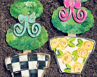 May include: Two painted wooden topiary trees in pots. The topiary on the left has a blue bow and a black and white checkered pot. The topiary on the right has a pink bow and a yellow pot with a floral pattern. The text "Sunflower Soul Art" is at the bottom of the image.