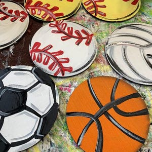 May include: Hand-painted wooden coasters featuring sports ball designs. Includes a yellow softball with red stitching, a white baseball with red stitching, a black and white soccer ball, an orange basketball, and a white volleyball.