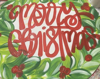 May include: Round wooden Christmas sign with a white background and red lettering that reads "Merry Christmas." The sign is surrounded by a green wreath with red berries.