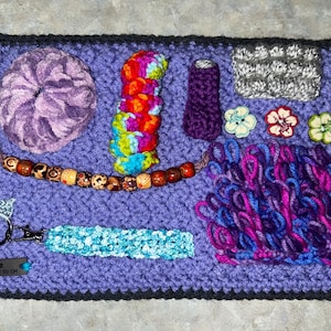 Puede incluir: Un tablero sensorial morado tejido a ganchillo con varios elementos texturizados. Incluye un gran remolino, una hebra de cuentas, un llavero y una pulsera. Otros elementos incluyen una pila de anillos de colores, un pequeño tubo morado, un cuadrado gris y botones en forma de flor.
