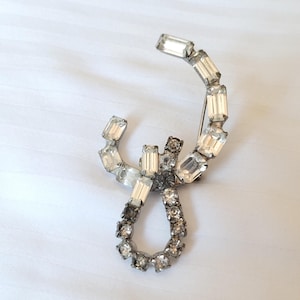 May include: Vintage silver-toned brooch with clear and smoky rhinestones. The brooch features a curved design with rectangular and round stones, creating a sparkling, elegant accessory. The pin is set against a white background.